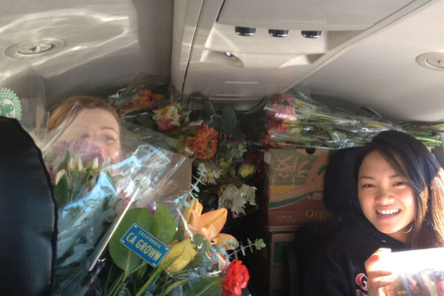 Campus Kitchen volunteers smile surrounded by flowers and food inside a vehicle.